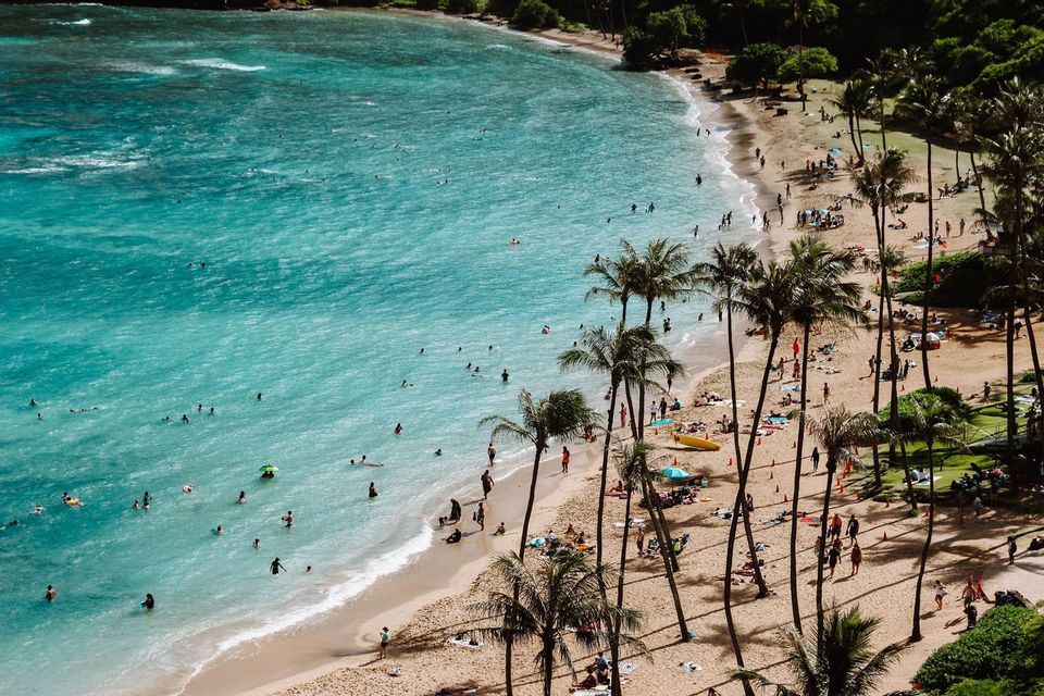 Ein Blick von oben auf einen belebten Tropenstrand mit Menschen, die in türkisfarbenem Wasser schwimmen und sich am Sandstrand unter Palmen entspannen.