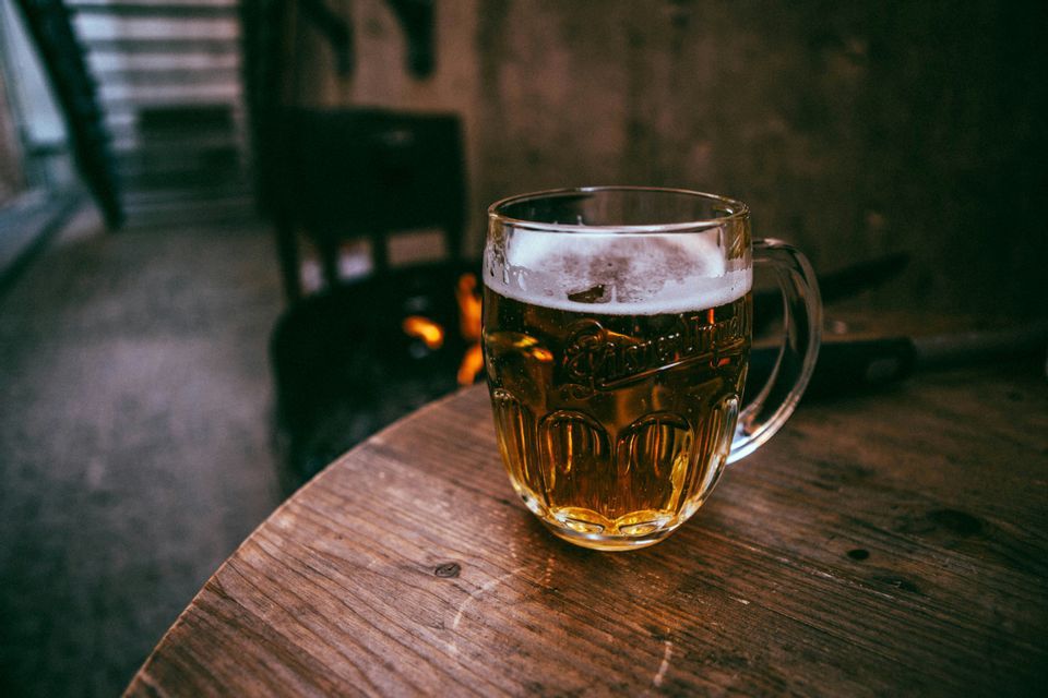 Un boccale di birra di vetro con una schiuma densa si trova su un tavolo di legno, illuminato da un fuoco scoppiettante in sottofondo.