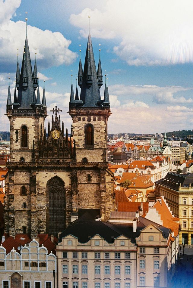 The twin spires of a Gothic church rise above the red rooftops of a historic city under a partly cloudy sky.