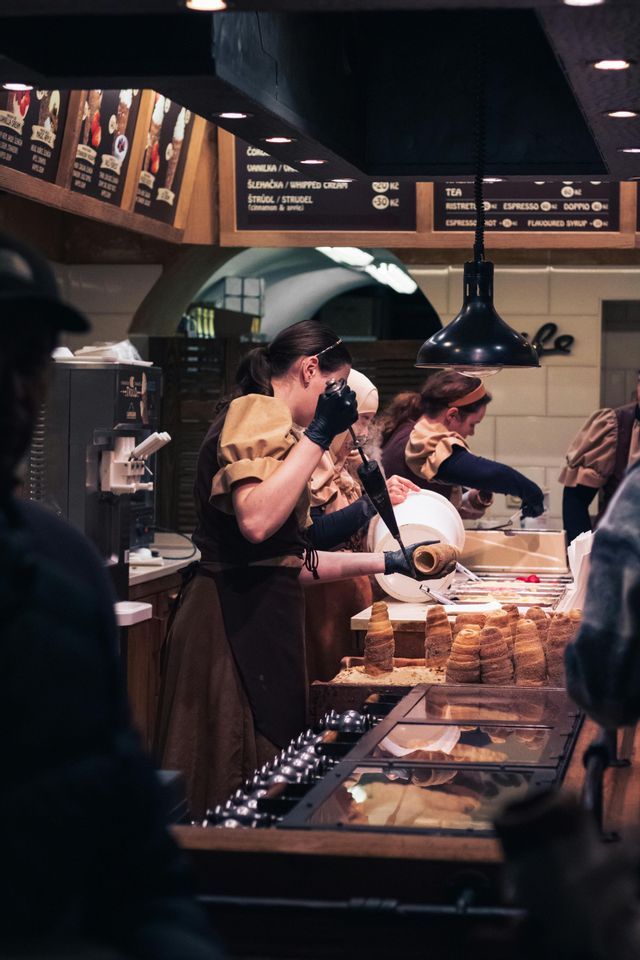 A woman in a uniform and black gloves fills a chimney cake pastry with a piping bag at a busy bakery counter.