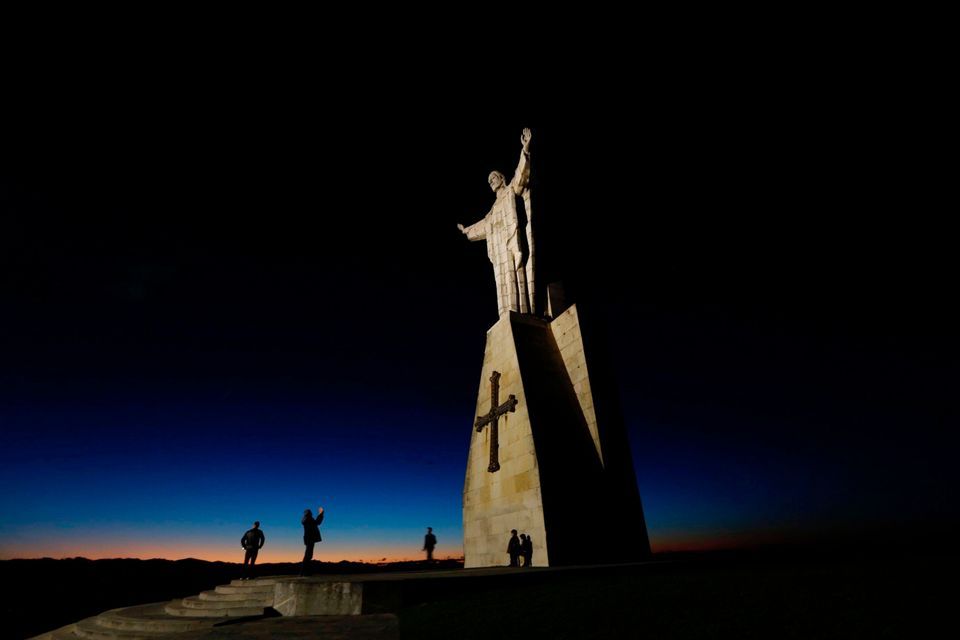 Una gran estatua iluminada se alza sobre un pedestal contra un cielo crepuscular, con las siluetas de varias personas en su base.