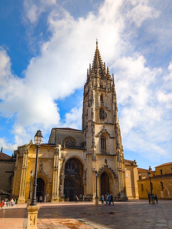 Una gran catedral gótica con una alta aguja se alza en una plaza pavimentada bajo un cielo azul con nubes blancas.