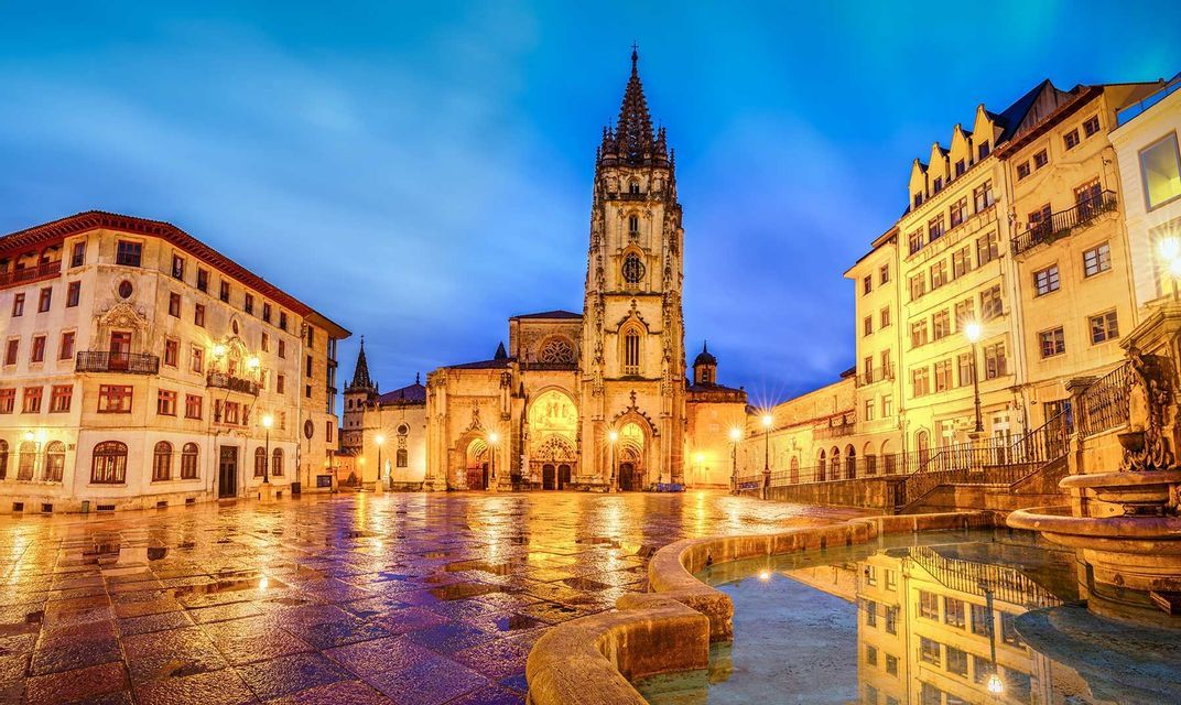 Una catedral gótica iluminada se alza en una plaza de la ciudad al anochecer, sus luces reflejándose en el suelo adoquinado mojado.