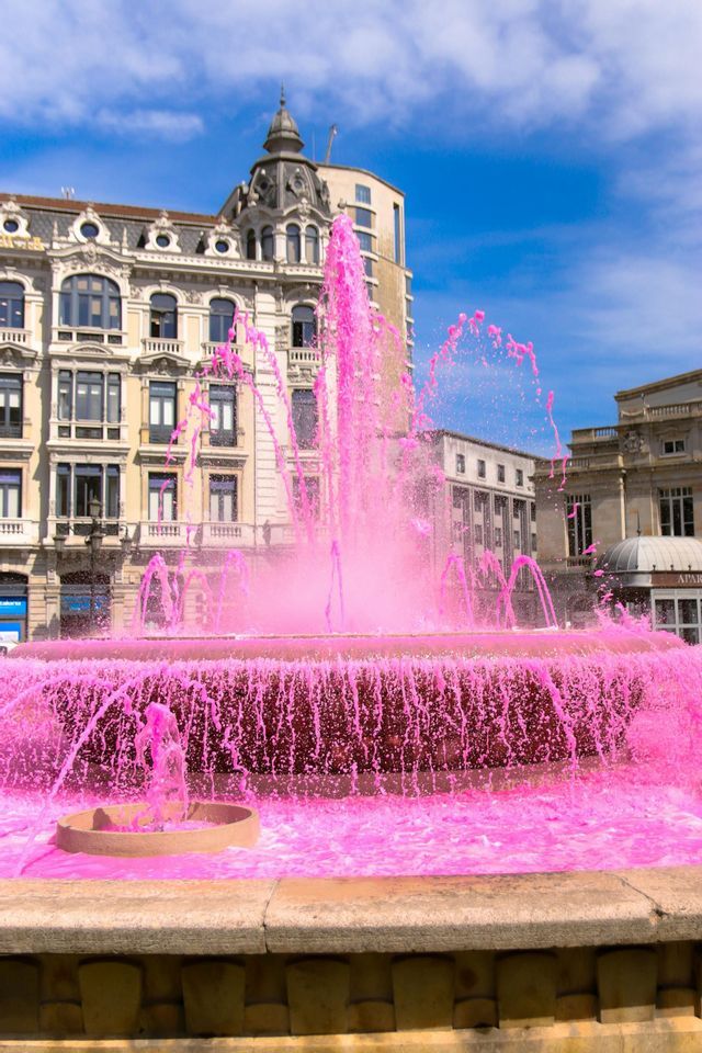 Una gran fuente rocía agua rosa vibrante en una plaza de la ciudad frente a edificios ornamentados bajo un cielo azul.