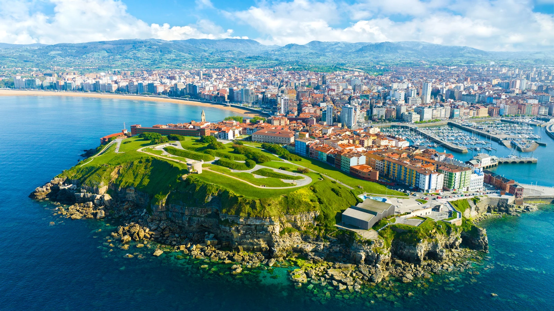 Vista aérea de una ciudad en una península verde y rocosa, con una larga playa de arena y un puerto deportivo en la costa.