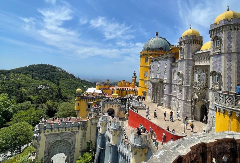 Un vivace palazzo giallo e grigio si erge su una lussureggiante collina verde, con turisti che passeggiano lungo le sue terrazze e cortili sotto un cielo azzurro.