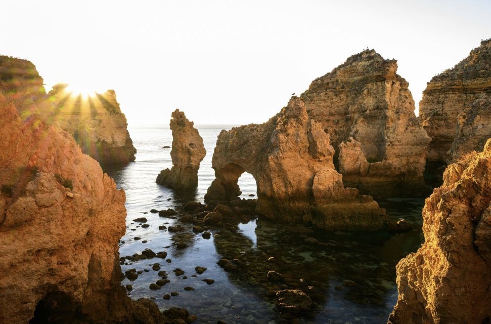 Il sole fa capolino da dietro grandi formazioni rocciose, incluso un arco naturale, che si ergono nelle calme acque dell'oceano.