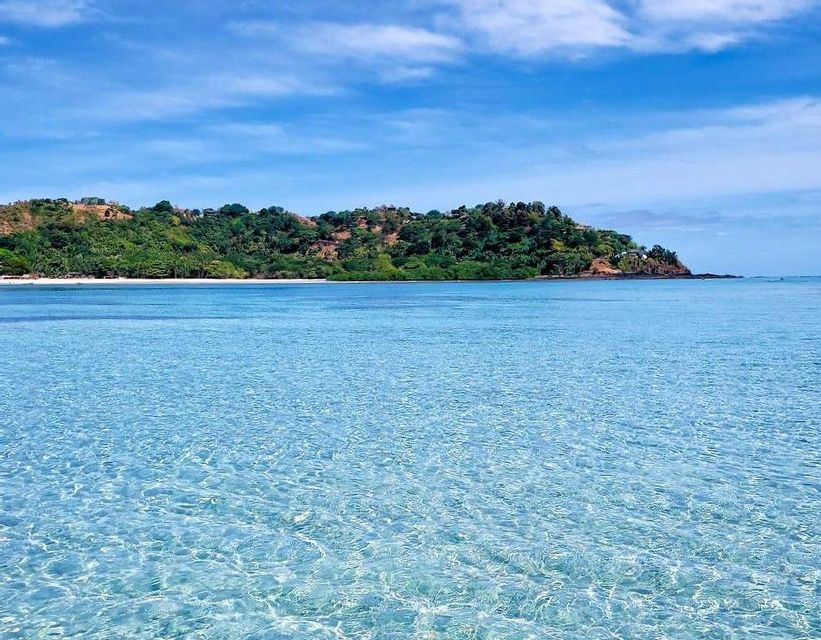 Acqua cristallina color turchese luccica davanti a un'isola verde lussureggiante con una piccola spiaggia di sabbia bianca sotto un cielo blu.