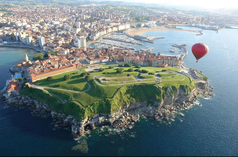 Un globo aerostático rojo flota sobre una ciudad costera con una península verde y acantilada, un puerto deportivo y una playa.