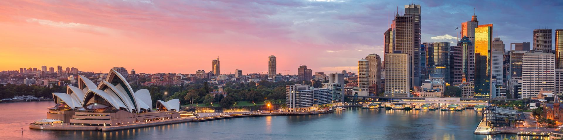 Una vista panorámica del horizonte de una ciudad con una ópera en el puerto, iluminada por un colorido atardecer.