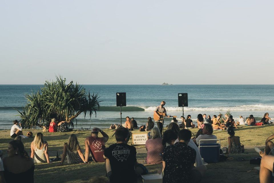 Un músico toca una guitarra acústica para una multitud sentada en una colina de hierba con vistas al océano.