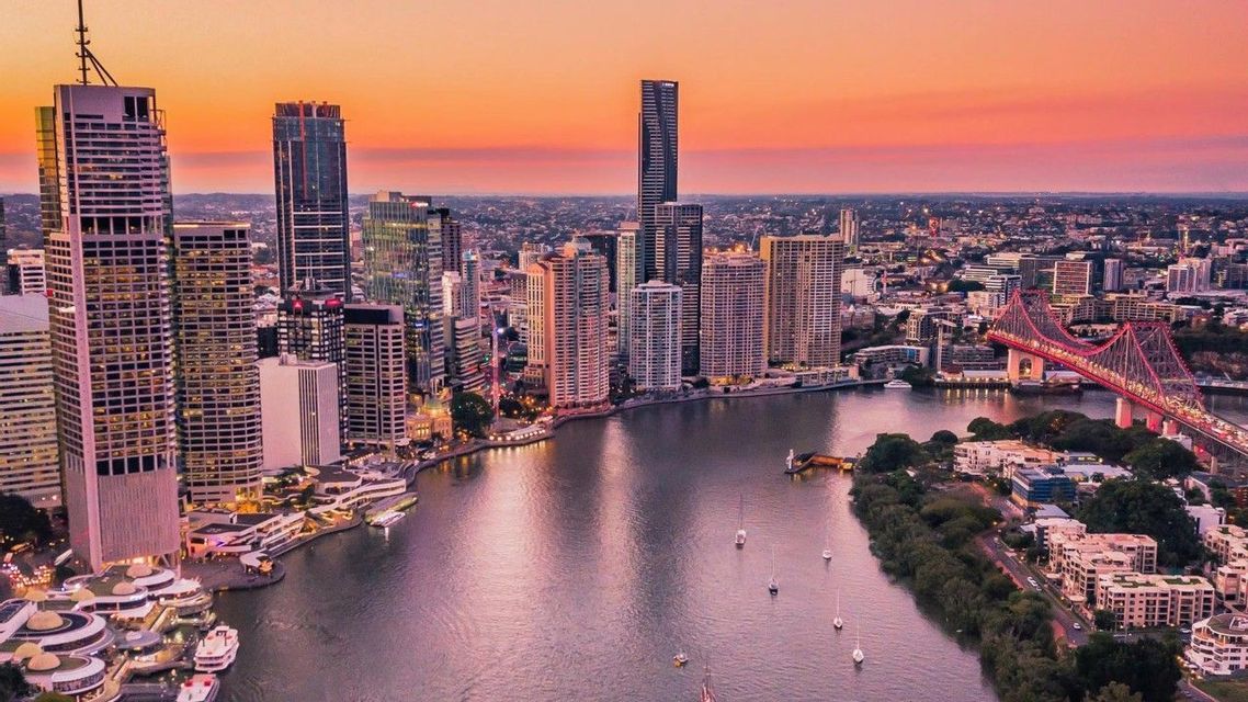 Una vista aérea de un paisaje urbano moderno con rascacielos a lo largo de un río serpenteante, un puente iluminado y veleros bajo un cielo al atardecer.