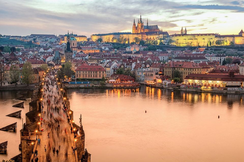 An illuminated stone bridge crowded with people crosses a river, leading to a historic cityscape with a castle on a hill at dusk.