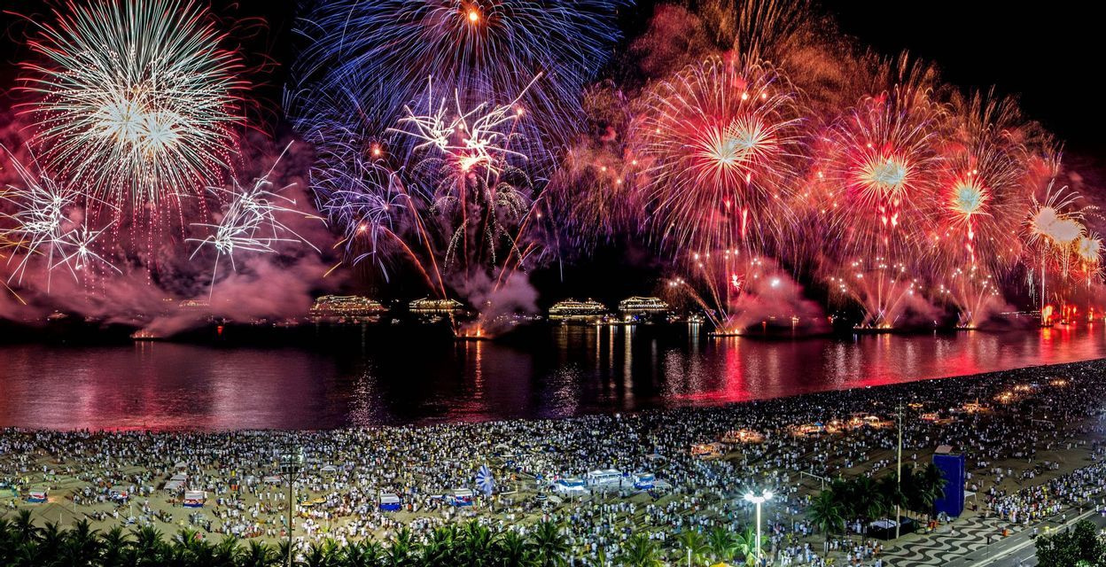 Buntes Feuerwerk über einem belebten Strand und einer Bucht mit mehreren beleuchteten Schiffen bei Nacht.