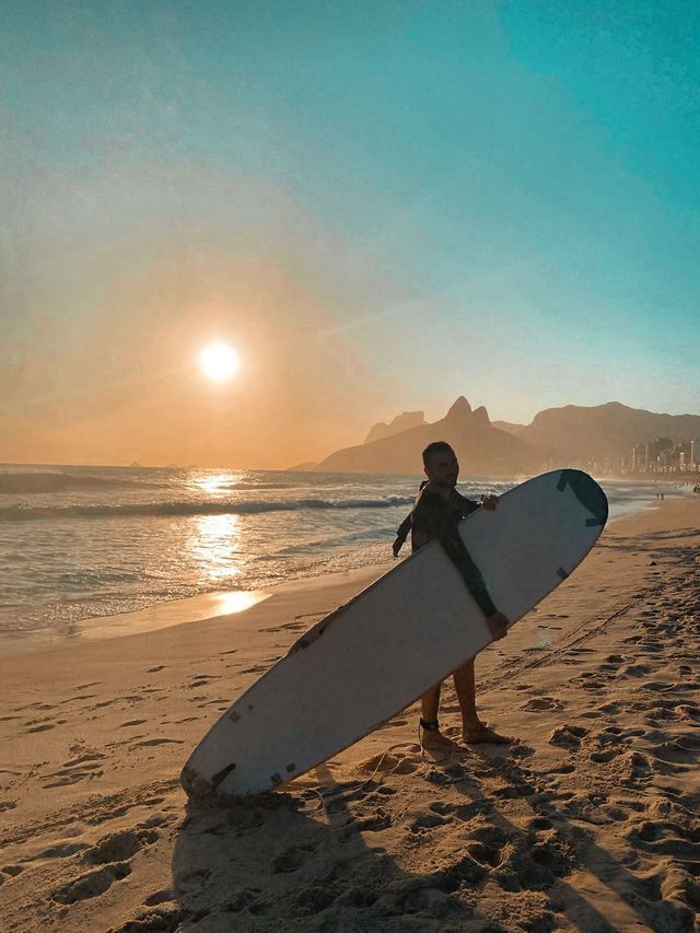 Un homme en combinaison de plongée tient une planche de surf debout sur une plage de sable au coucher du soleil, avec des montagnes et une ville en arrière-plan.