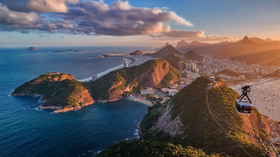 Una vista aerea da una funivia mostra una città costiera incastonata tra montagne verdi e l'oceano al tramonto.