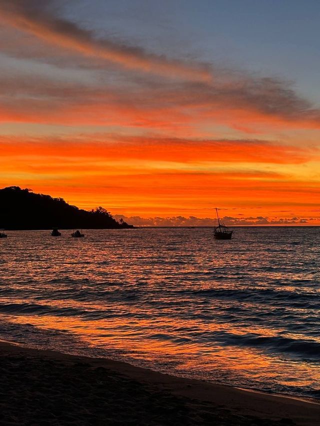 L'oceano riflette un tramonto vibrante arancione e rosso, mentre diverse barche sono stagliate contro una costa scura.