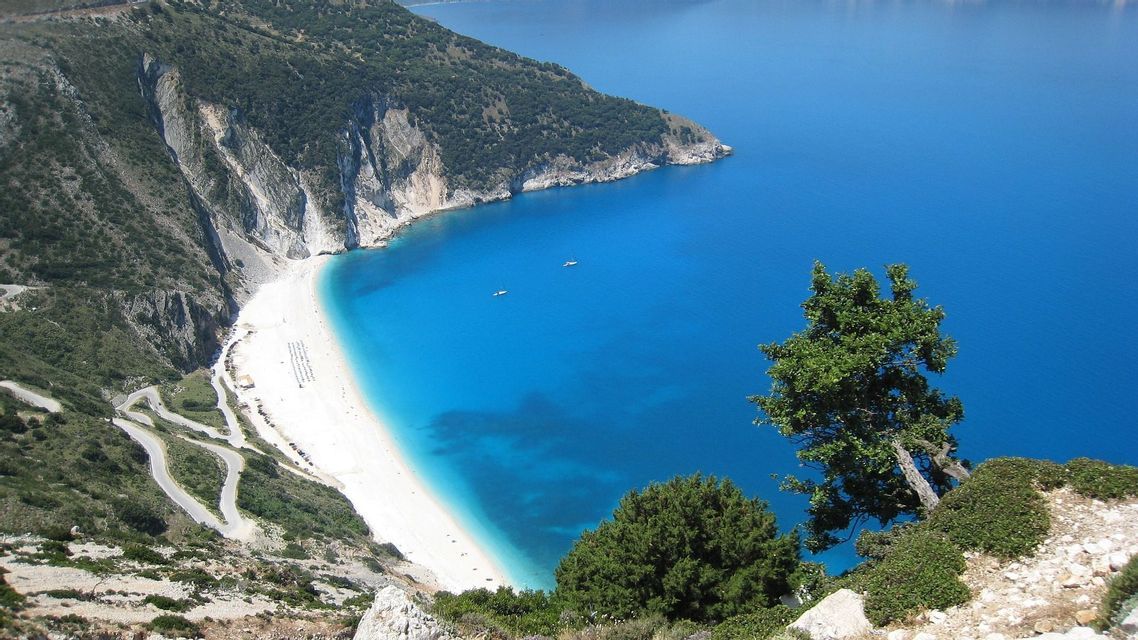 Vista dall'alto di una cala di sabbia bianca con scogliere a picco, una strada costiera tortuosa e due barche sul mare turchese.