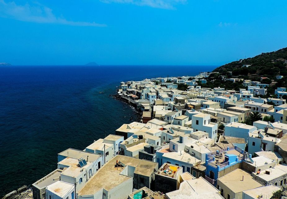 Veduta aerea di un borgo costiero con case bianche e tetti piani che si affaccia sul mare blu profondo.