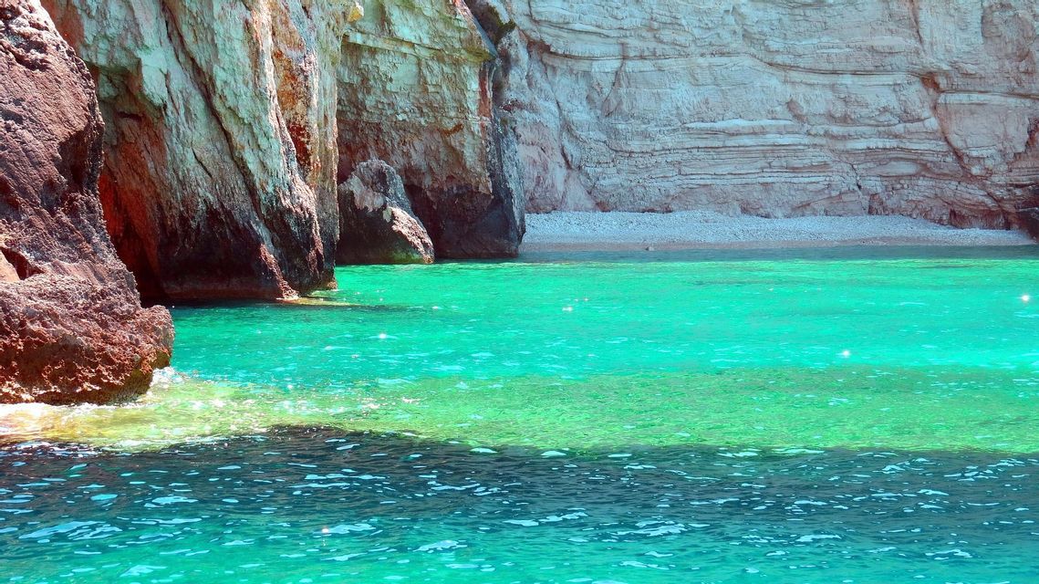 Acque turchesi e vibranti del mare incontrano la base di alte scogliere rocciose che costeggiano una spiaggia di ciottoli appartata.