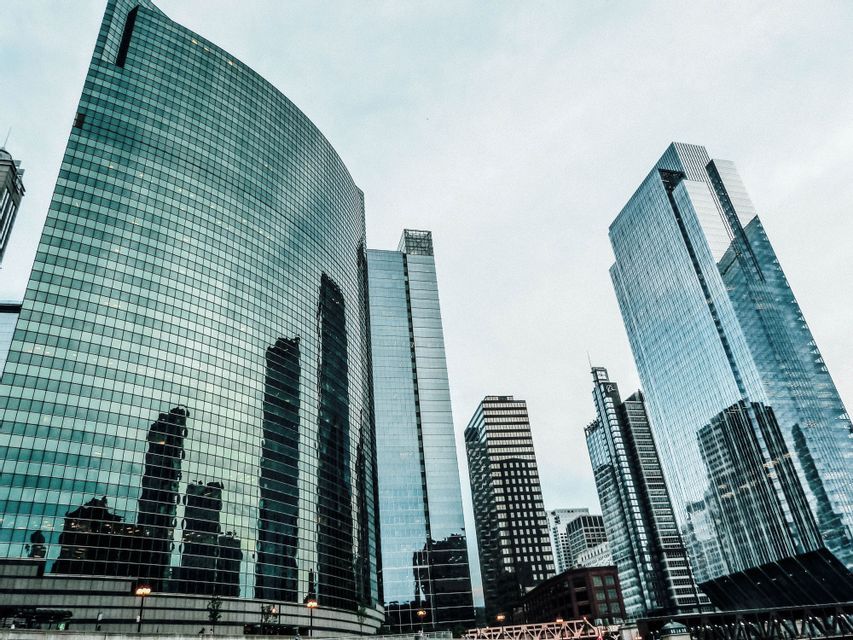 Vista dal basso di imponenti grattacieli di vetro, con una facciata curva che riflette altri edifici contro un cielo coperto.