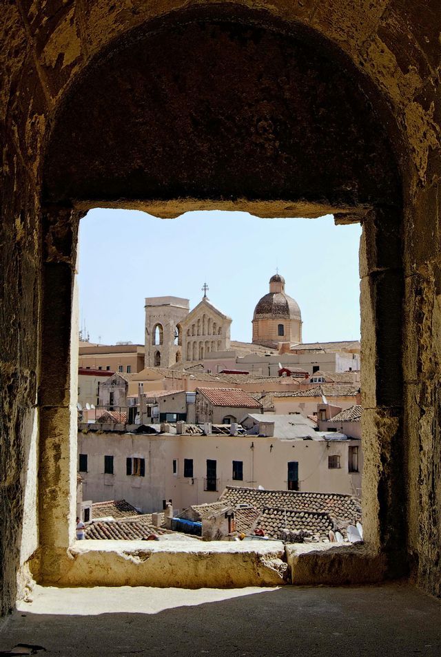 Un paisaje urbano soleado con tejados de terracota y una cúpula de iglesia, visto a través del marco de un arco de piedra.