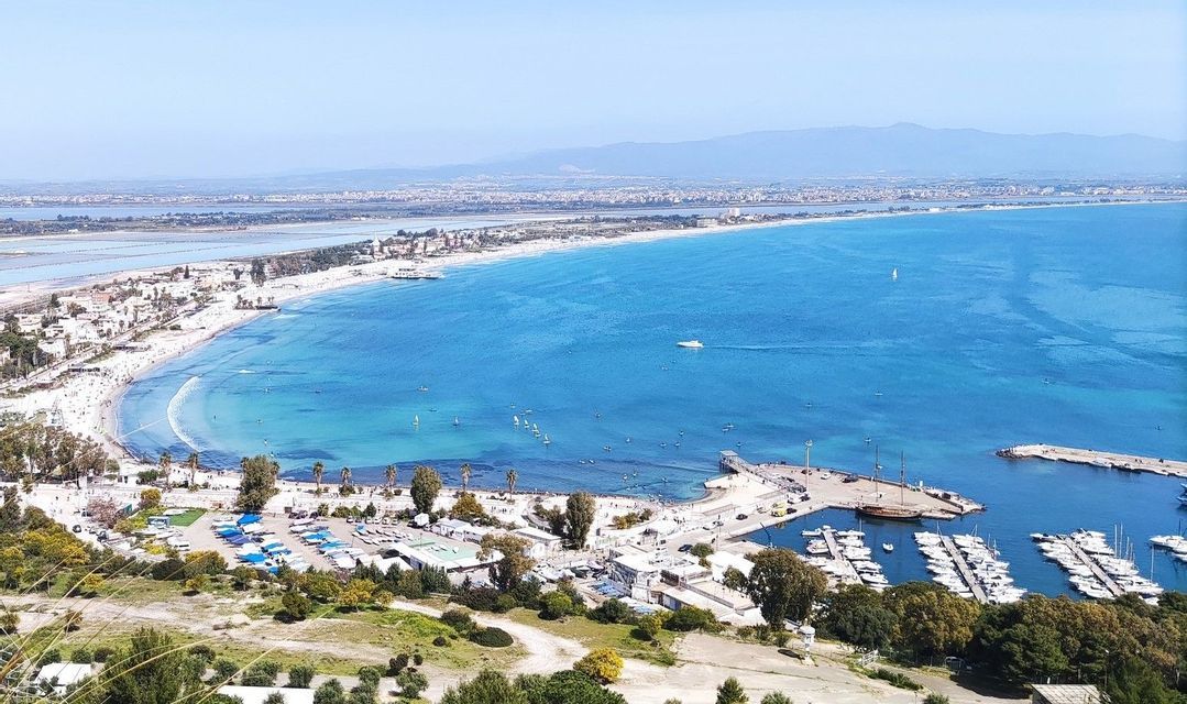 Una vista aérea de un paisaje costero con una playa de arena blanca curvada, un puerto deportivo y una bahía azul brillante con veleros.