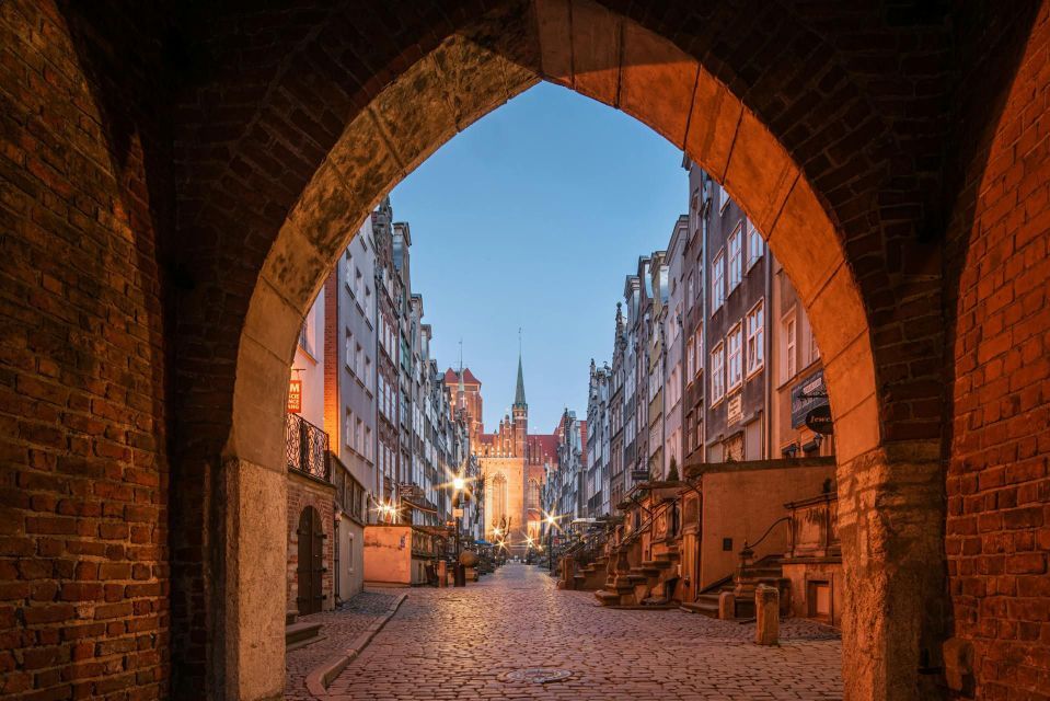 Eine gepflasterte Straße, gesäumt von historischen Gebäuden, unter einem warmen Backsteinbogen in der Dämmerung gesehen.