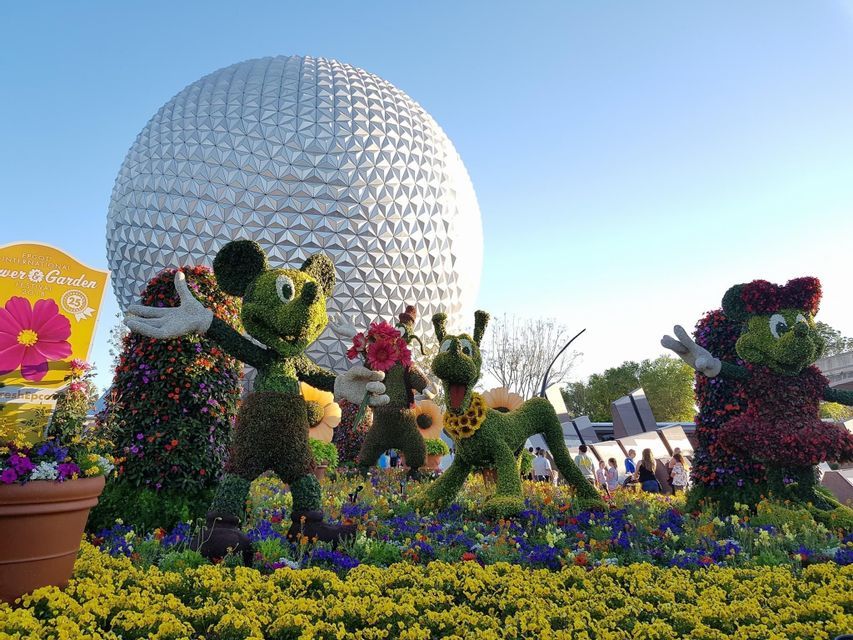 Des topiaires de Mickey Mouse, Pluto et Minnie Mouse se dressent dans un parterre de fleurs éclatant, avec une grande sphère géodésique en arrière-plan.