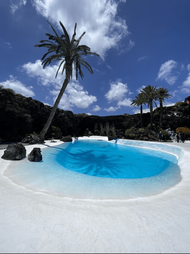 Una piscina azzurra luminosa in stile laguna è circondata da terreno bianco, palme e roccia vulcanica scura sotto un cielo nuvoloso.