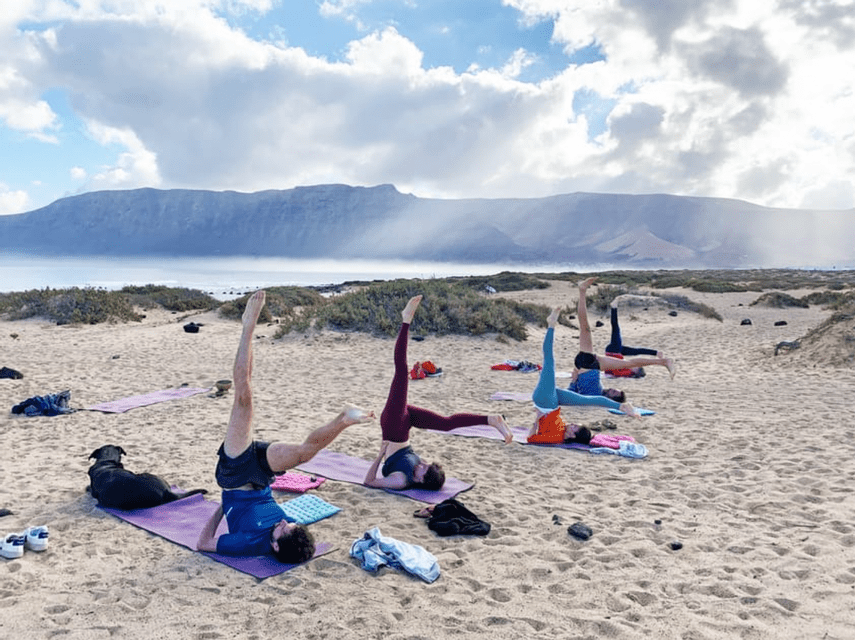 Un gruppo WeRoad pratica yoga su tappetini in una spiaggia sabbiosa, con un cane nelle vicinanze e montagne sullo sfondo.