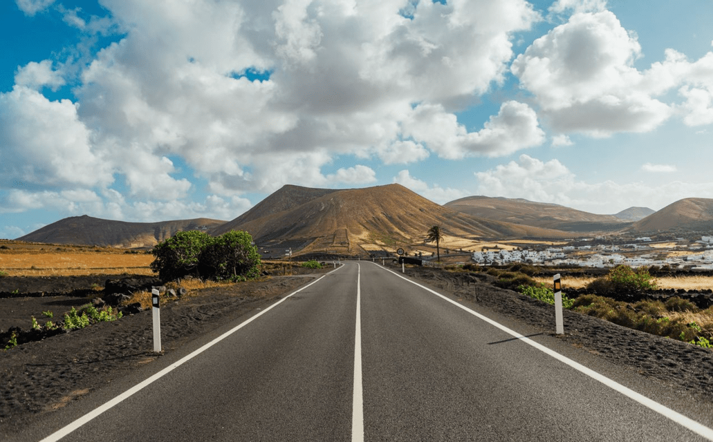 Una strada asfaltata attraversa un paesaggio vulcanico verso le montagne e una piccola città sotto un cielo azzurro nuvoloso.