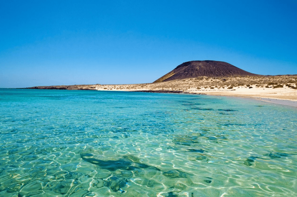 Acque turchesi limpide lambiscono una spiaggia sabbiosa con un vulcano dormiente sullo sfondo sotto un cielo azzurro senza nuvole.
