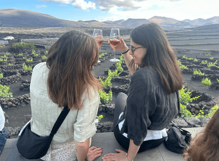 Due donne di un viaggio di gruppo WeRoad brindano con bicchieri di vino, con vista su un vigneto vulcanico e montagne in lontananza.