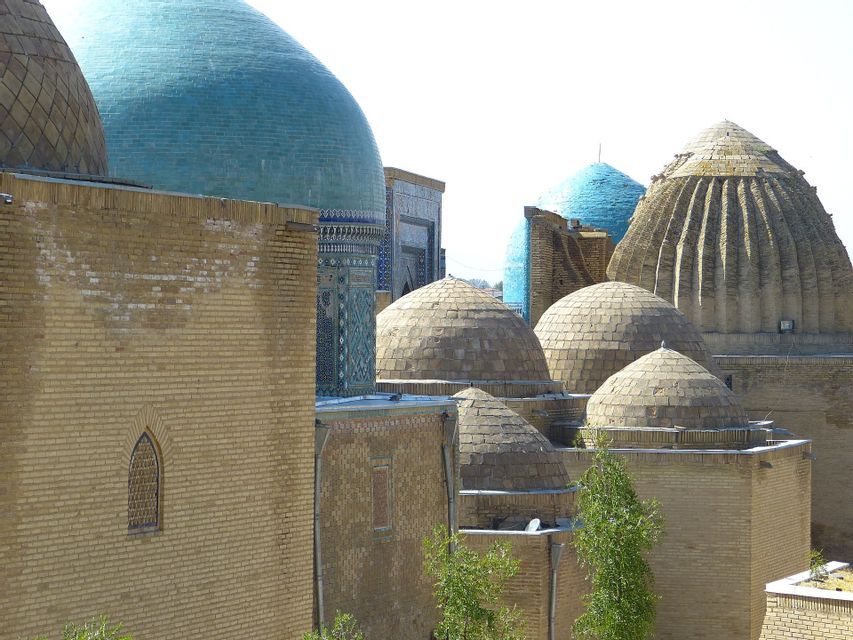 Una collezione di cupole in pietra e piastrelle blu brillante sopra edifici in mattoni color sabbia sotto un cielo sereno.