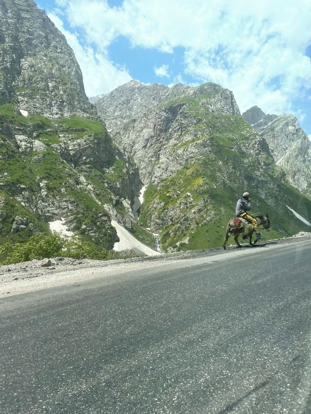 Una persona su un asino viaggia su una strada accanto a montagne ripide e rocciose con chiazze di erba verde e neve.