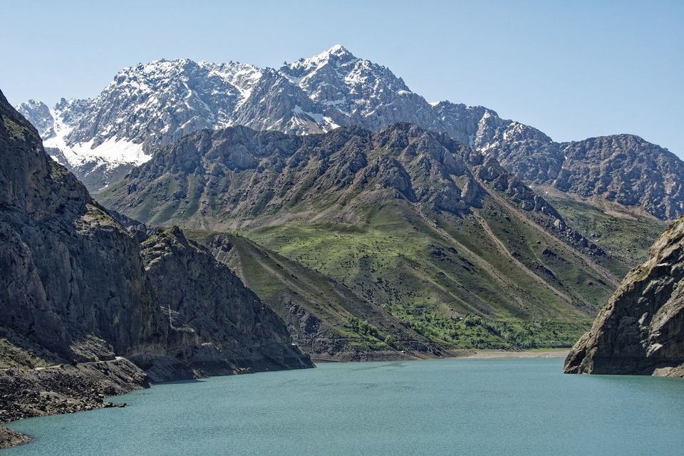 Un lago turchese in una valle circondata da ripidi pendii verdi e montagne rocciose, con cime innevate sullo sfondo.