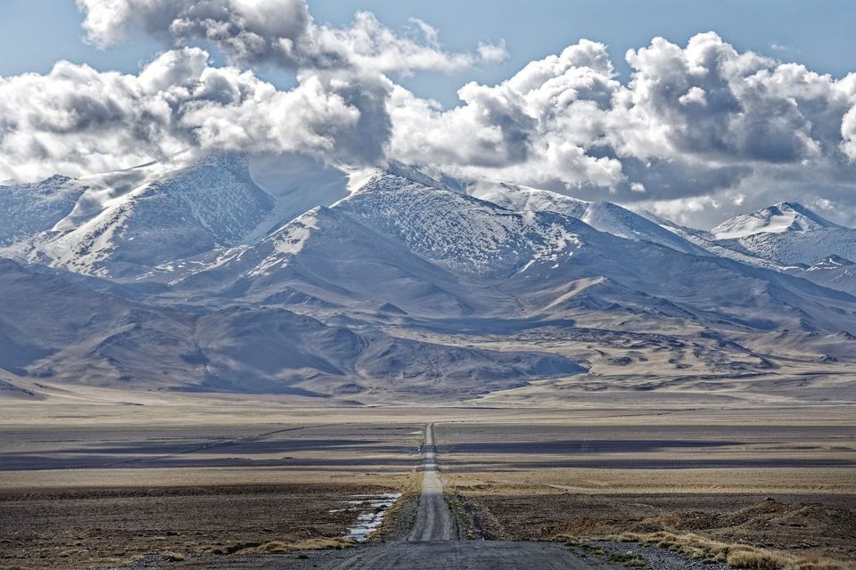Una lunga e dritta strada sterrata si estende attraverso una vasta pianura arida verso una catena montuosa innevata sotto un cielo nuvoloso.
