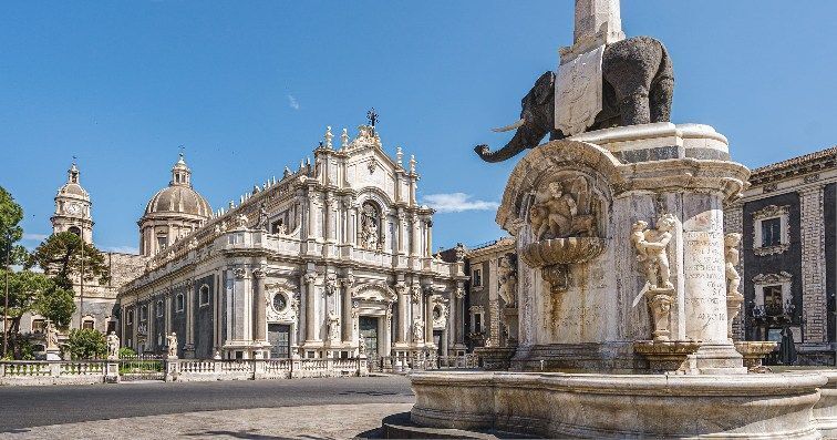 Una fontana in pietra con una statua di elefante in una grande piazza pavimentata, con una cattedrale ornata sullo sfondo sotto un cielo sereno.