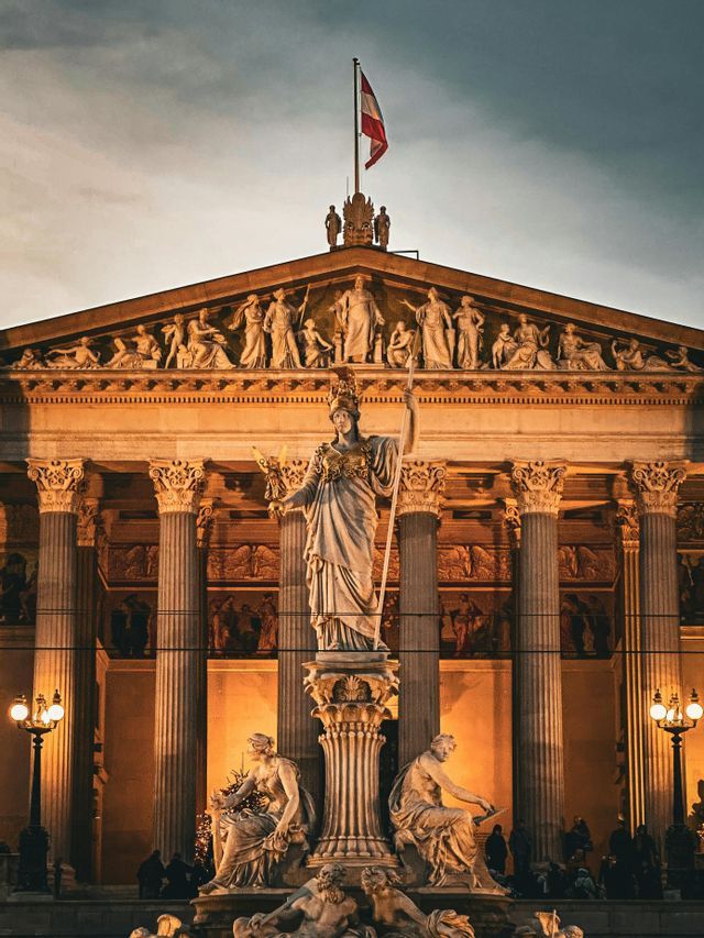 Una statua di una guerriera con una lancia davanti a un edificio classico illuminato con colonne e una bandiera austriaca