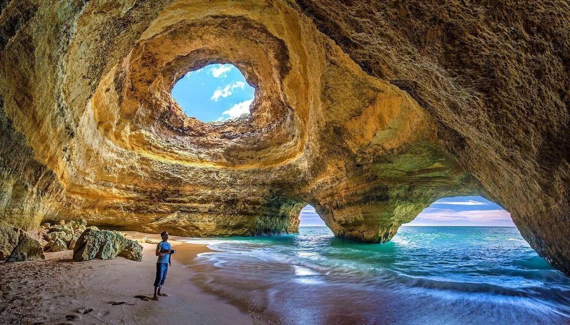 A person stands on a sandy beach inside a large sea cave with an archway to the ocean and a circular opening in the ceiling.