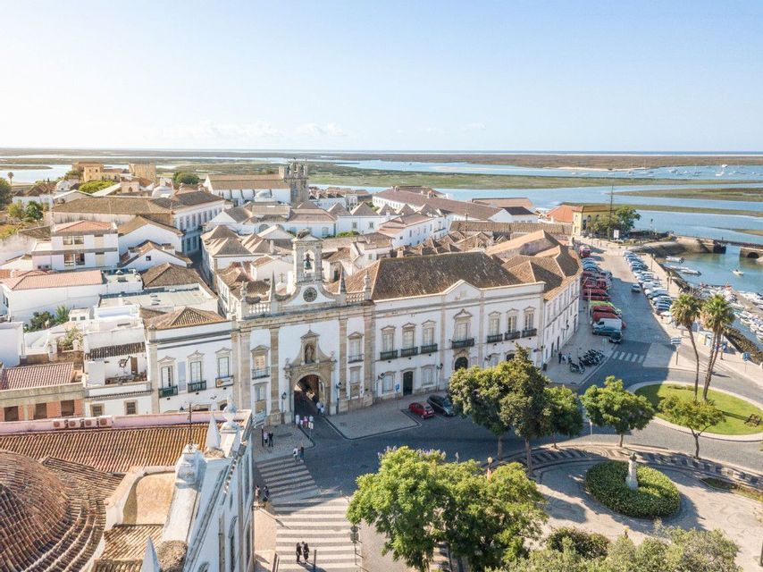 Aerial view of a coastal town with white buildings, an ornate central archway, and a harbor with boats on a sunny day.