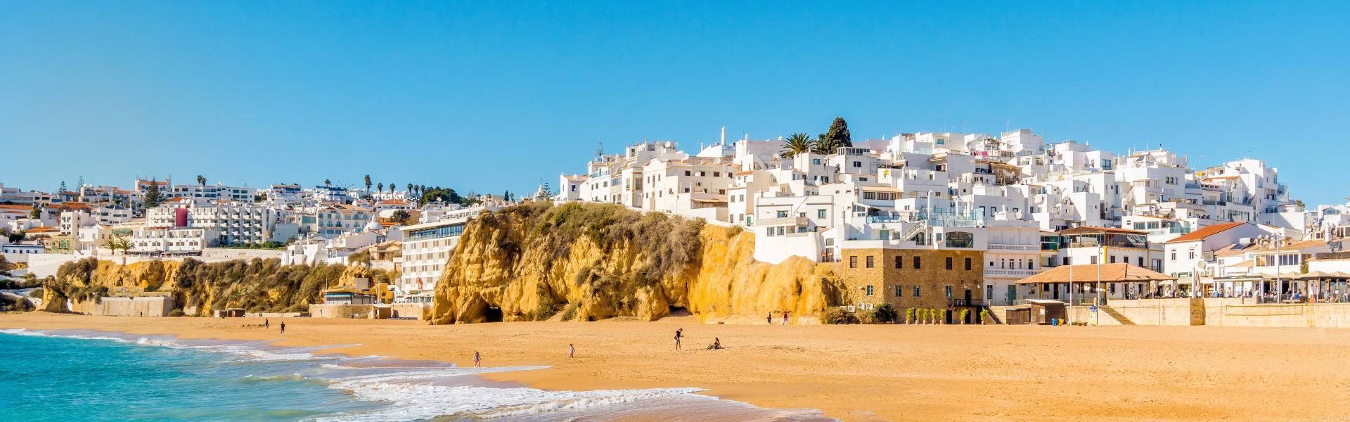 A panoramic view of a coastal town with whitewashed buildings on a cliff above a golden sand beach and the ocean.