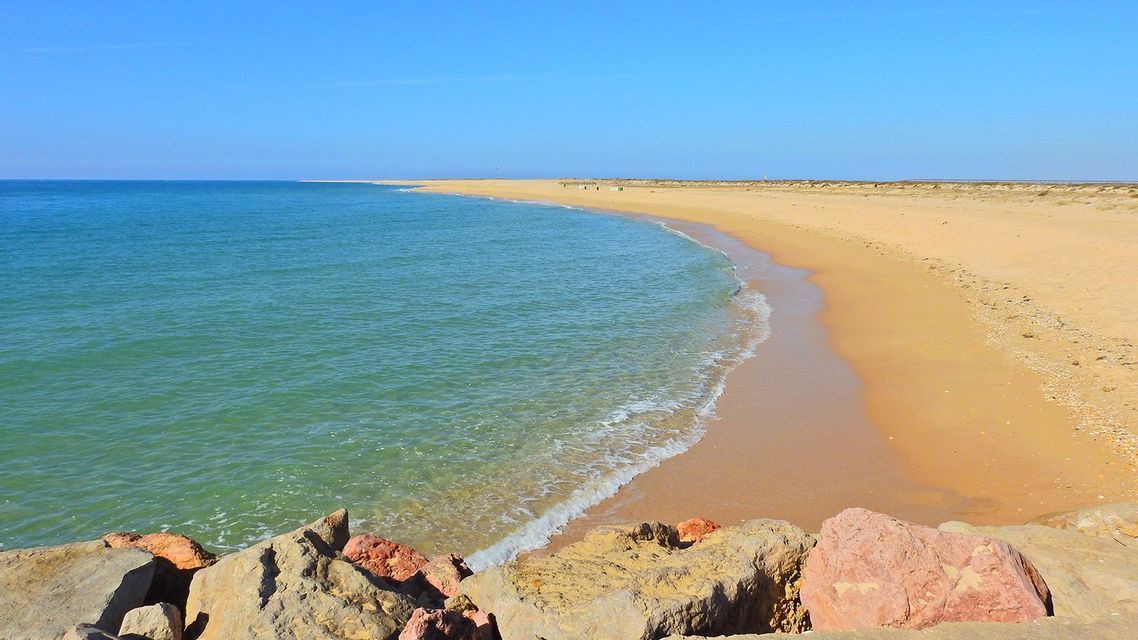 A view from a rocky outcrop shows calm turquoise water lapping against a long sandy beach under a clear blue sky.
