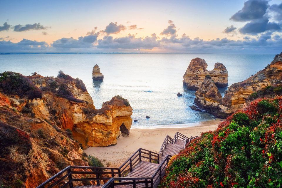 Wooden stairs lead down a vegetated cliff to a sandy beach with sea stacks in the ocean at sunrise.