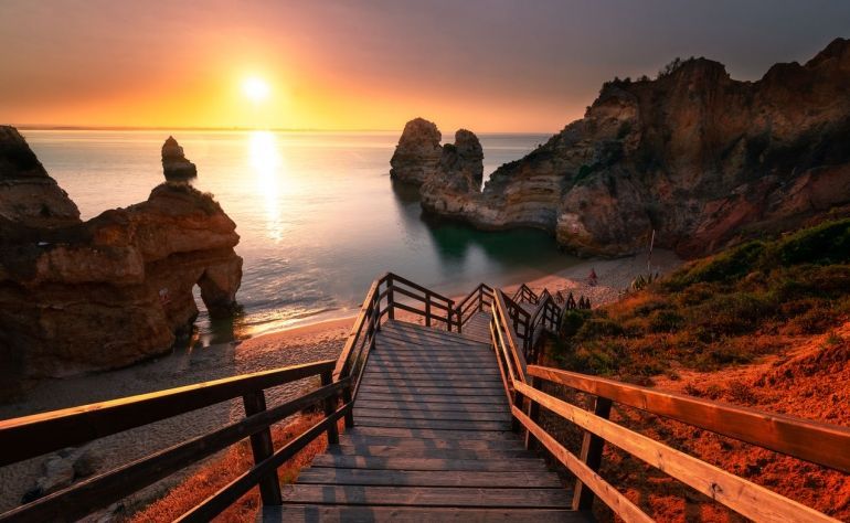 A wooden staircase leads down to a sandy beach cove with large rock formations in the ocean at sunset.