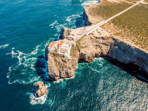 Aerial view of a white lighthouse with a red roof on a rugged cliff, overlooking a deep blue ocean with crashing waves.