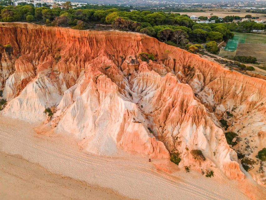 An aerial view of eroded red and white cliffs towering over a sandy beach, with a dense green forest at the top.