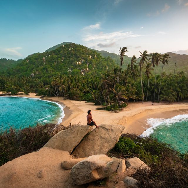 Ein Mann sitzt auf einem großen Felsen und überblickt zwei tropische Strände mit türkisfarbenem Wasser, Palmen und einem üppig grünen Hügel.