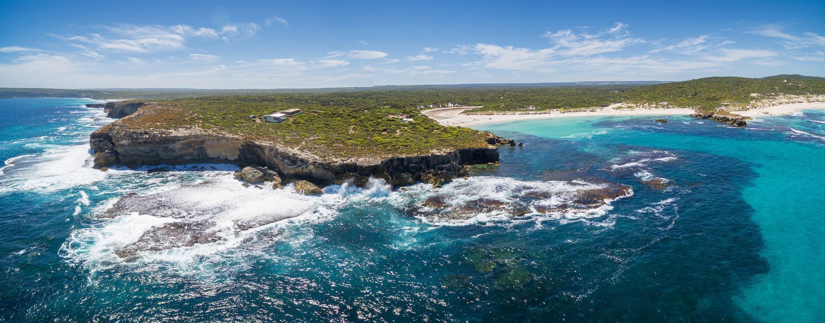 An aerial view of a rugged, green coastline with a rocky headland, a white sand beach, and turquoise ocean water under a blue sky.